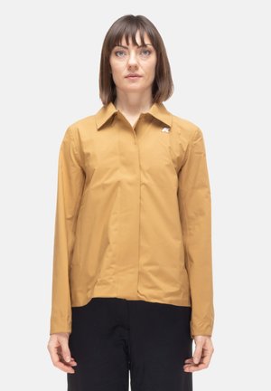 Donna con capelli castani corti che indossa una giacca a maniche lunghe color senape e pantaloni neri, in piedi davanti a uno sfondo chiaro.