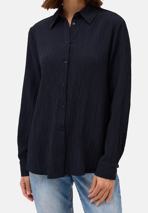 Femme portant une chemise noire texturée à manches longues avec boutons, associée à un jean bleu clair, montrée du cou aux hanches.
