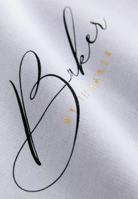 Biela látka s čiernym kurzívnym nápisom "Baker" a zlatým nápisom "by Ted Baker" pod ním. Textúra pôsobí hladko so slabým leskom.