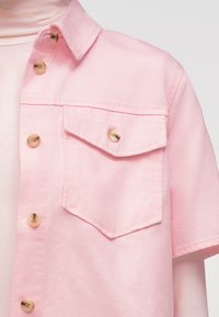 Ροζ ανοιχτό ροζ πουκάμισο με κοντά μανίκια και πατιλέτα στην τσέπη στο στήθος που ασφαλίζει με κουμπί, που φοριέται πάνω από ανοιχτό ροζ μπλουζάκι με ζιβάγκο.