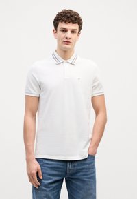 Biele polo tričko s šedým pruhovaným golierom, krátkymi rukávmi a malým logom na hrudi. Nosené s modrými džínsami.