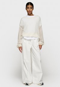 Pull bianco oversize, in tessuto strutturato, con maniche lunghe, indossato sopra una camicia bianca. Abbinato a pantaloni bianchi a gamba larga e sneaker grigie.