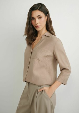 Kobieta o ciemnych, falowanych włosach nosi beżową zapinaną na guziki bluzkę z rękawami do połowy przedramienia oraz beżowe spodnie z wysokim stanem, jedna ręka w kieszeni, neutralne tło.