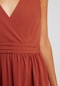 Rukávový šat rustové barvy s výstřihem do V, vypasovaným pasem a nabíranou sukni na osobě se světlou hnědou pletí.