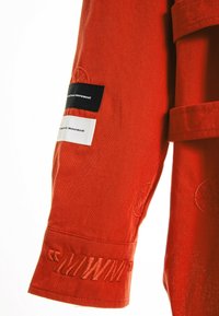 Manga de chaqueta de mezclilla roja con etiquetas en blanco y negro, acentos bordados y tela texturizada. Los detalles visibles en el puño resaltan el diseño.