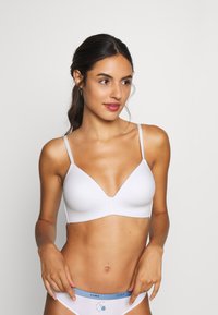 Bralette blanche avec des fines bretelles et un décollet�é en V profond, présentant une texture lisse. Porté avec des culottes blanches ayant une ceinture bleue.