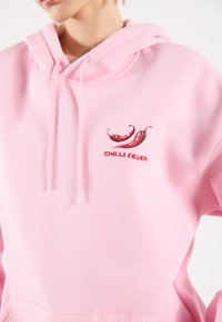 Svetlorúžová mikina s klokaním vreckom, šnúrkami a grafickým motívom červenej chili papričky s textom "CHILI FEVER." Mäkkučký materiál, ležérny strih.