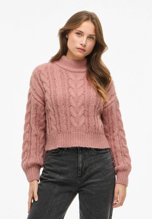 Pull en tricot rose avec un col montant, présentant un motif en tricot torsadé et des poignets côtelés. Longueur cropped associé à un jean noir taille haute.
