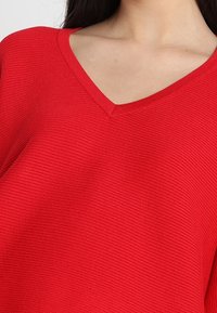 Czerwony, prążkowany sweter z V-dekoltem i luźnym krojem. Materiał ma gładką teksturę i subtelny pionowy wzór.