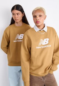 Dva pojedinca nose prevelike, bež dukserice s bijelim logom "New Balance". Jedan ima košulju s kragnom ispod; drugi ima dugu kosu.