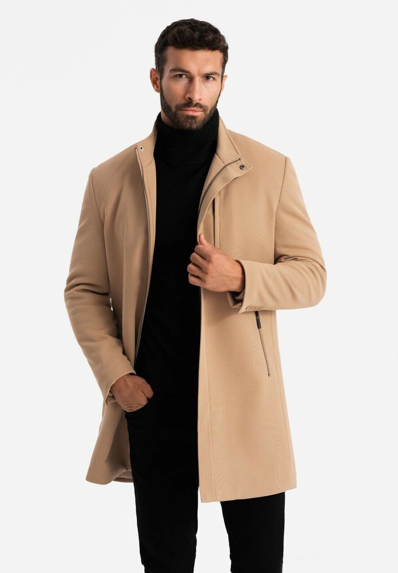 Cappotto beige con superficie texturizzata, colletto alto e chiusura con zip. Indossato sopra un dolcevita nero, con tasche laterali e vestibilità su misura.