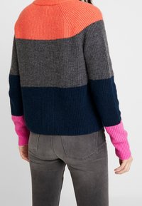 Objemný svetr s ribbovanou texturou, zdobený pruhovanými panely v korálové, šedé, námořnické a růžové barvě, kombinovaný se šedými kalhotami.