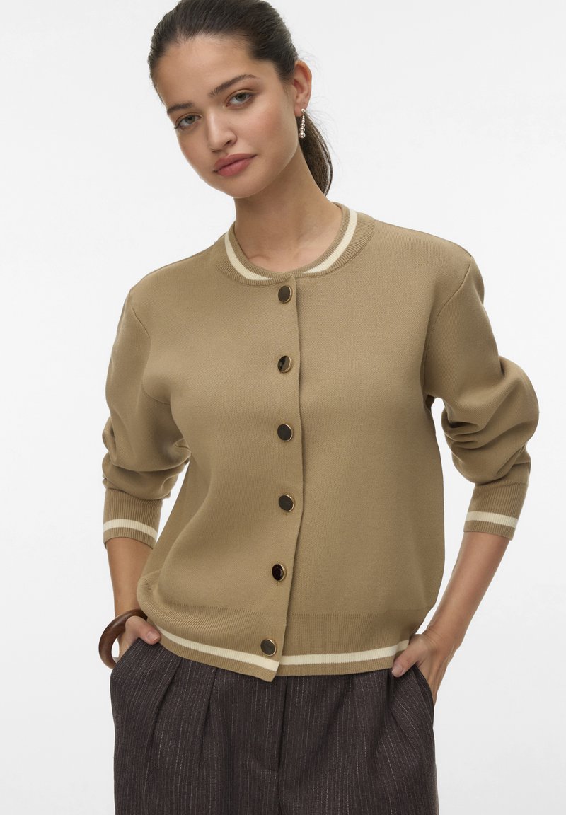 Khaki gebreide cardigan met een ronde halslijn, gouden knopen en crèmekleurige strepen bij de mouwen en onderkant, gecombineerd met donkere geplooide broeken.