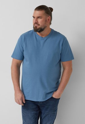 Erwachsener Mann mit Bart und zurückgebundenem Haar, der ein blaues Kurzarmhemd und Jeans trägt, mit einer Hand in der Tasche, schaut nach links.