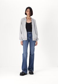 Γκρι ζακέτα με φερμουάρ, εφαρμοστό μαύρο μπλουζάκι, ψηλόμεσα μπλε τζιν με φαρδιές άκρες και μαύρες πλατφόρμες παπούτσια. Απλό φόντο.