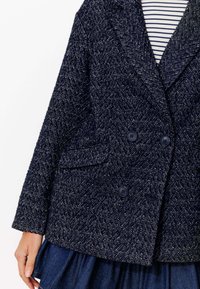 Manteau double boutonnage bleu marine texturé avec un motif en tweed, deux poches avant et des boutons noirs, porté avec une chemise rayée en dessous.