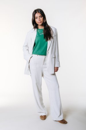 Costume rayé gris clair avec pantalon ample et blazer oversize, associé à un t-shirt vert. Porte des chaussures couleur caramel et un collier chaîne simple.