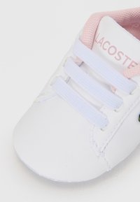Baskets blanches pour enfant en matériau synthétique, avec des accents roses, trois perforations pour les lacets avec des lacets blancs, et le logo Lacoste sur la languette.