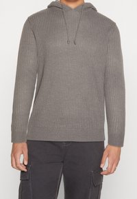 Grau gestrickte Kapuzenjacke mit gerippter Textur, langen Ärmeln, verstellbarem Kordelzug und lässigem Schnitt. Kombiniert mit dunklen Cargo-Hosen.