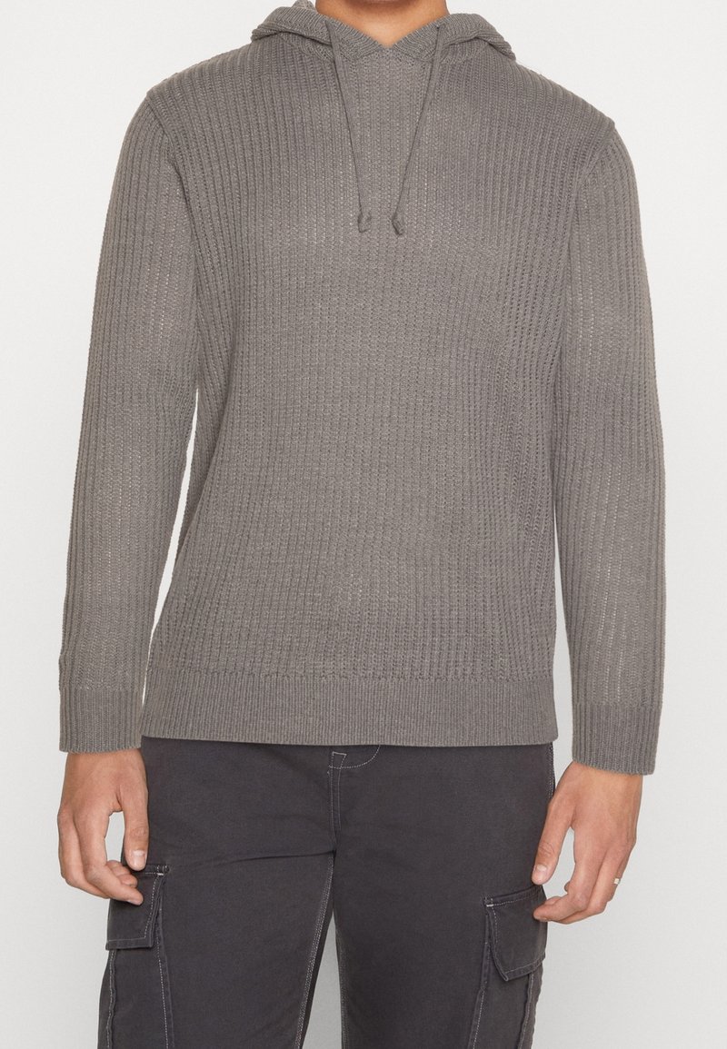 Grau gestrickte Kapuzenjacke mit gerippter Textur, langen Ärmeln, verstellbarem Kordelzug und lässigem Schnitt. Kombiniert mit dunklen Cargo-Hosen.
