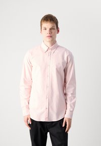 Lys rosa langermet skjorte med knapper og krage, med et lite brodert logo på brystet og en glatt bomullstekstur.