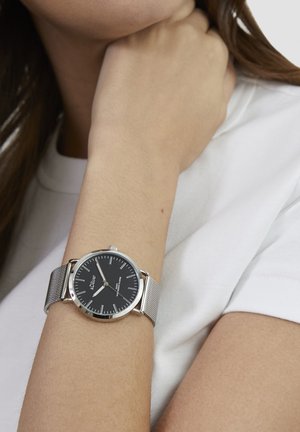 Runde Uhr mit schwarzem Zifferblatt, silbernen Stundenmarkierungen und einem silbernen Mesh-Armband. Getragen an einem nackten Handgelenk vor einem weißen Hemd.