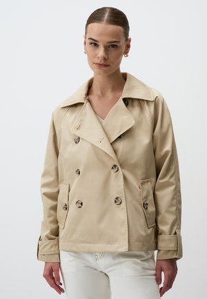 Giacca beige doppiopetto realizzata in materiale lucido, presenta grandi bottoni, un ampio colletto e tasche laterali. Texture liscia.