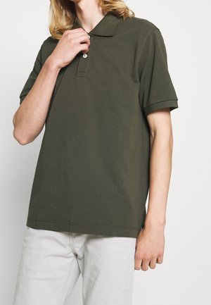 Ung person iført olivengrøn kortærmet poloshirt og lyse bukser, justerer skjortekraven med den ene hånd.