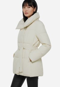 Giacca esterna bianca puffa con colletto alto, vita con cintura e tasche laterali. Design trapuntato testurizzato con finitura liscia.