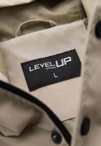 Beige Jackeninnenseite mit einem schwarzen Etikett, auf dem "LEVEL UP ombre" und die Größe "L" steht, sichtbarer Reißverschluss und schwarze Taste auf dem Stoff.