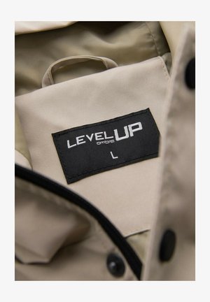 Interno del giubbotto beige con un'etichetta nera che riporta la scritta "LEVEL UP ombre" e la taglia "L", con cerniera visibile e bottone nero sul tessuto.