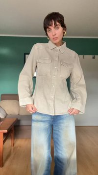 Camisa de mezclilla beige de manga larga, con cierre de botones, dos bolsillos en el pecho y dobladillo curvado, combinada con jeans de pierna ancha azul claro.