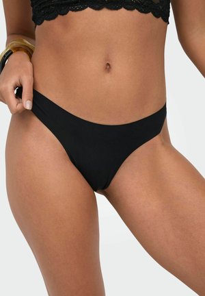 Woman wearing black bikini bottom and black lace crop top, adjusting bikini bottom on her hip against a plain background.