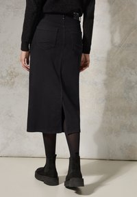 Falda midi de mezclilla negra con una abertura en la parte trasera, dos bolsillos traseros y una textura suave, combinada con botas de tobillo negras y medias opacas.