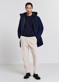Cappotto con cappuccio blu navy, maglione blu navy a texture, pantaloni plissettati color crema e stivaletti neri. Ambientato su uno sfondo chiaro e uniforme.