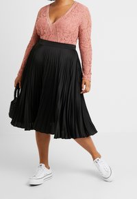Top in pizzo a maniche lunghe di colore rosa cipria con scollo a V, abbinato a una gonna midi plissettata nera e sneakers bianche, mentre tiene una borsa nera con perline.