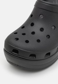 Gros plan d'un sabot en mousse noire avec des trous de ventilation et une semelle texturée sur fond blanc.