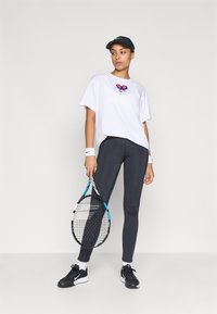 Λευκή μπλούζα με ροζ και μπλε γραφικά τένις, συνδυασμένη με σκούρες γκρι κολάν. Αξεσουάρ περιλαμβάνουν ένα μαύρο καπέλο και μαύρα αθλητικά παπούτσια, κρατώντας ένα ρακέτα τένις.