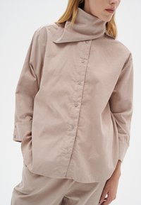 Lys beige skjorte med knapper og høj krave, lavet af tekstureret stof. Har lange ærmer og en afslappet pasform.