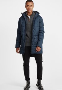 Jachetă puffer navy cu fermoar pe față, glugă ajustabilă, două buzunare în față și textură matlasată, purtată peste un pulover tricotat gri și pantaloni negri.