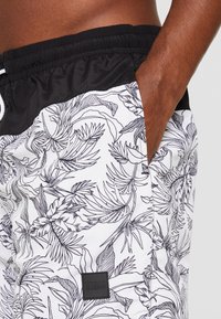 Sorte og hvite svømmeshorts med tropisk bladmønster, elastisk midjebånd, snor og en lapp med teksten "URBAN CLASSICS."
