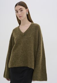 V nyakú, oversized pulóver olíva zöld színben, puha, bolyhos anyagból készült. Széles ujjakkal és laza szabással rendelkezik, amelyet egy fekete szoknyával párosítottak.