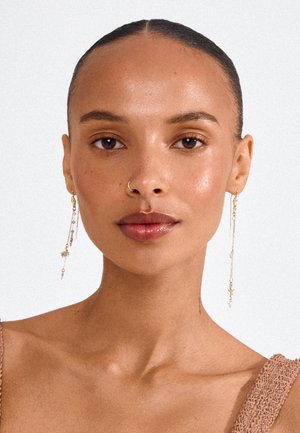 Mujer con piel suave y cabello peinado hacia atrás, que lleva pendientes largos y delicados de cadena dorada y un aro en la nariz, con un fondo liso.