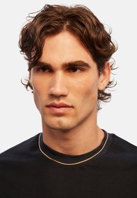 Guldtonet metal kæde halskæde båret over en mørk T-shirt med rund hals. Halskæden har et glat, buet design med en delikat tekstur.