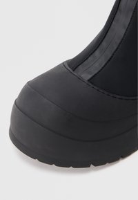 Černá nepromokavá bota s kulatou špičkou, hladkým gumovým materiálem, strukturovanou podrážkou a vertikálními švy na horní části.