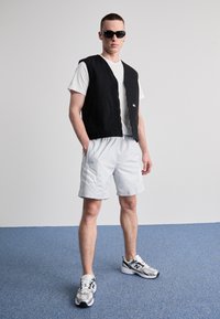 Colete preto sem mangas sobre uma t-shirt branca, calções cinzentos claros com riscas e ténis cinzentos. A pé sobre um chão revestido a carpete azul.