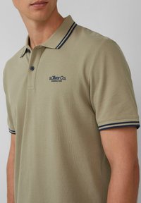 Khaki polo z teksturowanej tkaniny z granatowym kołnierzykiem w paski. Posiada małe granatowe logo i dwa guziki przy szyi.