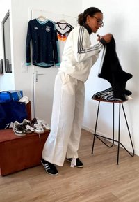 Persona con abbigliamento Adidas bianco che piega capi neri in una stanza con scarpe sportive e magliette della nazionale di calcio tedesca appese alla porta.