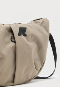 Μπεζ υφασμάτινη τσάντα με ρυθμιζόμενο μαύρο λουρί και ένα μαύρο στυλιζαρισμένο λογότυπο στο μπροστινό μέρος, παρουσιάζεται σε απλό ανοιχτόχρωμο φόντο.