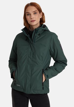 Vrouw draagt een donkergroene capuchonjas met ritsen en zakken aan de voorkant, handen in de zakken, staand tegen een effen lichte achtergrond.